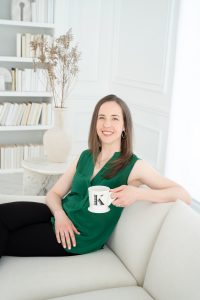Dr. Kate Whimster, ND sitting on an off-white couch holding a mug with the letter K on it and smiling.  She is wearing a green top and black pants.  In the background is a bookshelf full of books.
